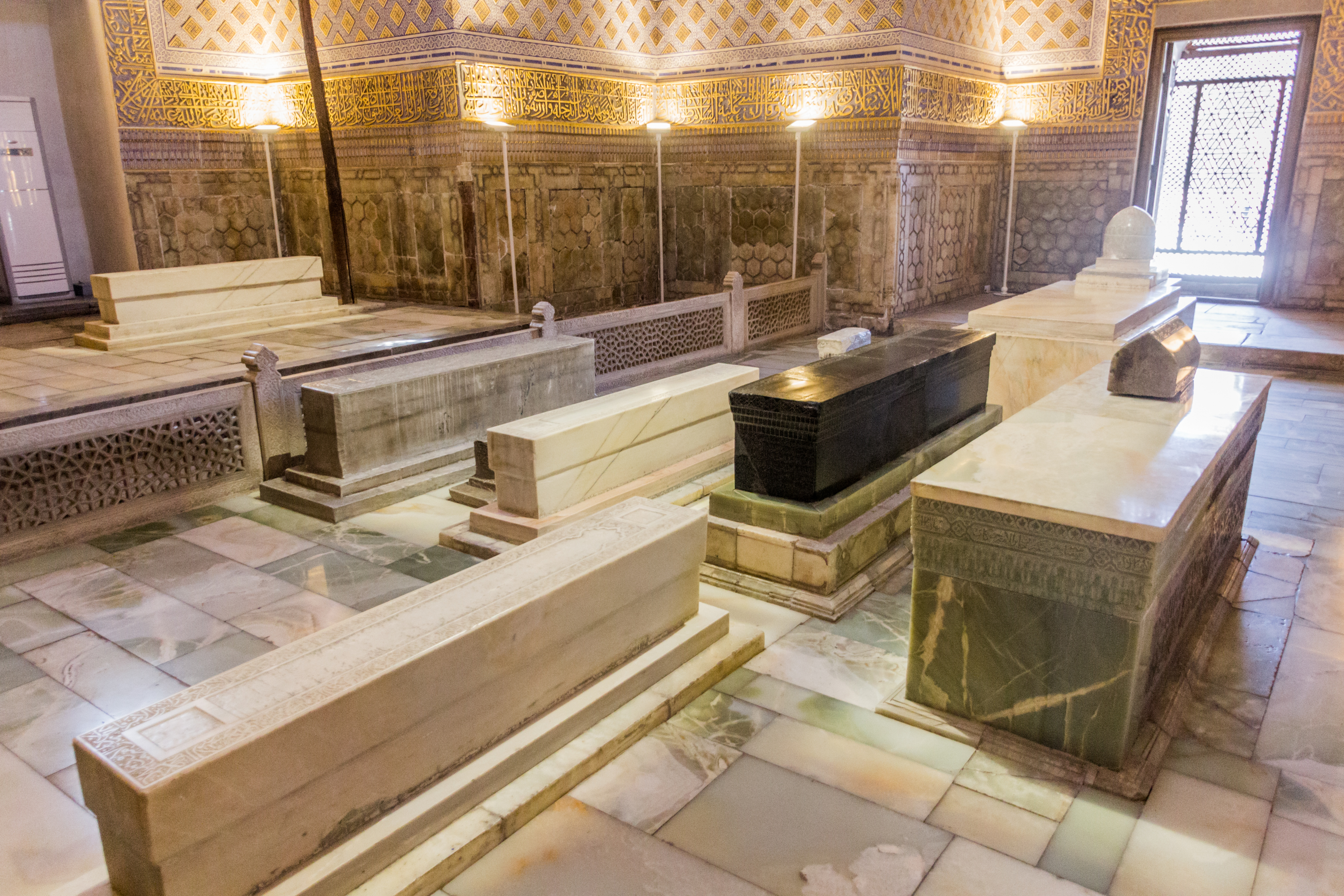 Interior of a highly decorated building with ornate carvings on the wall including the honeycomb pattern mentioned previously. A number of graves are in the foreground with the central one being topped with a black stone.