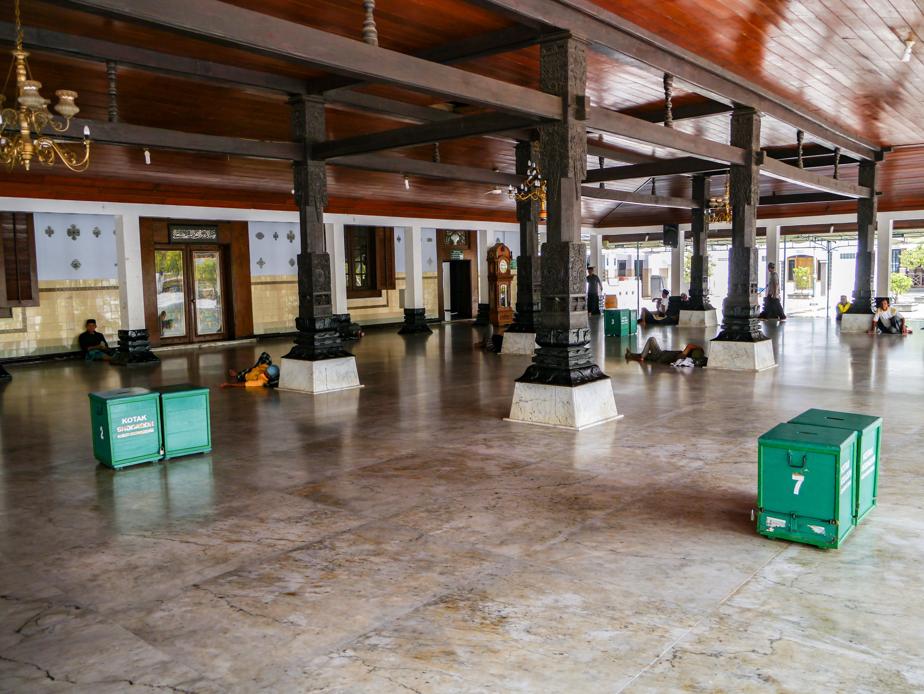 Courtyard with wooden pillars holding up a shining wooden ceiling. Some figures are seen sitting leaning against the pillars or relaxing on the floor.