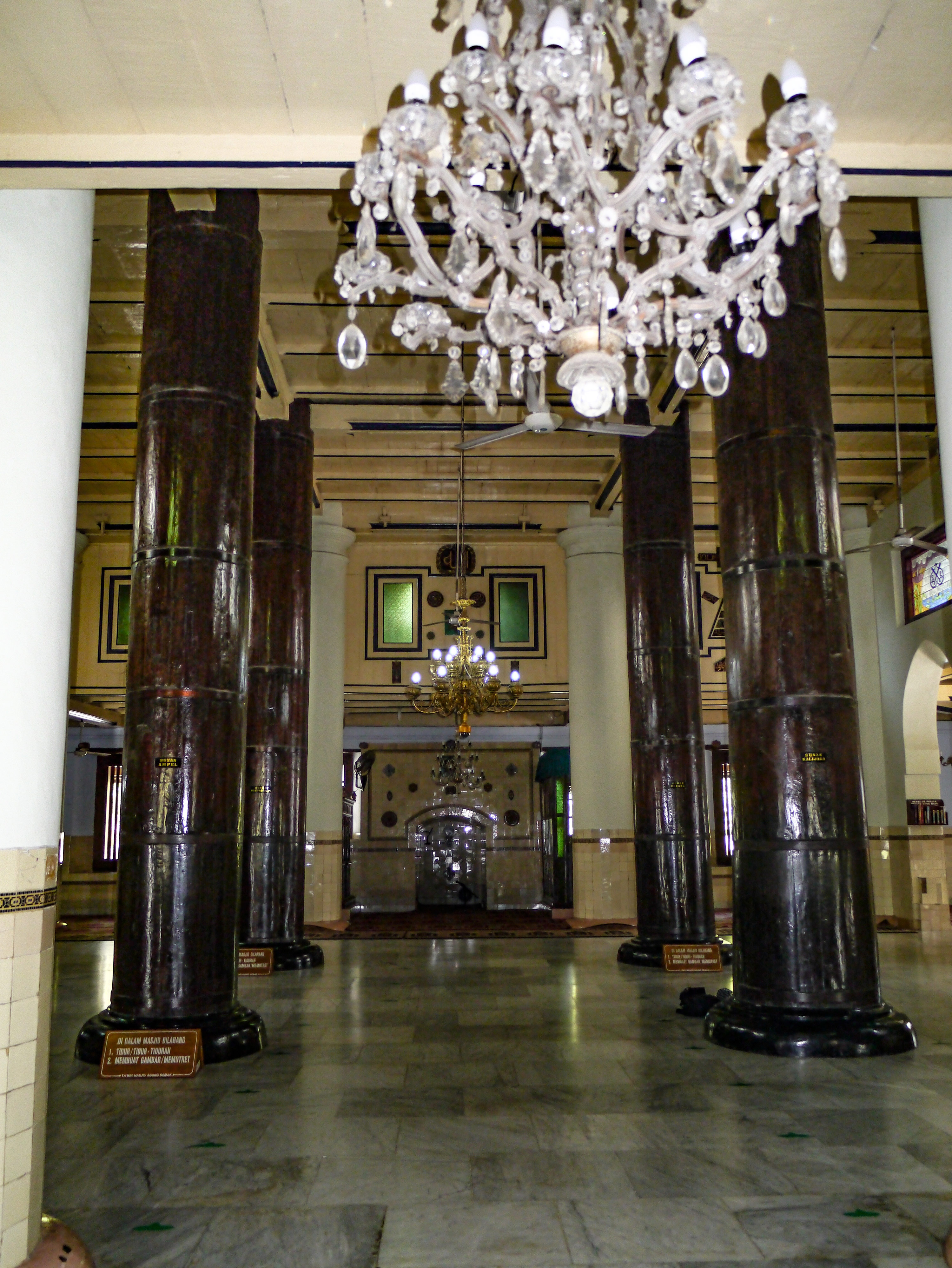 Four sizable wooden pillars in the mosque looking on to a prayer niche. Chandeliers are seen hanging along the ceiling, with one in the immediate foreground.