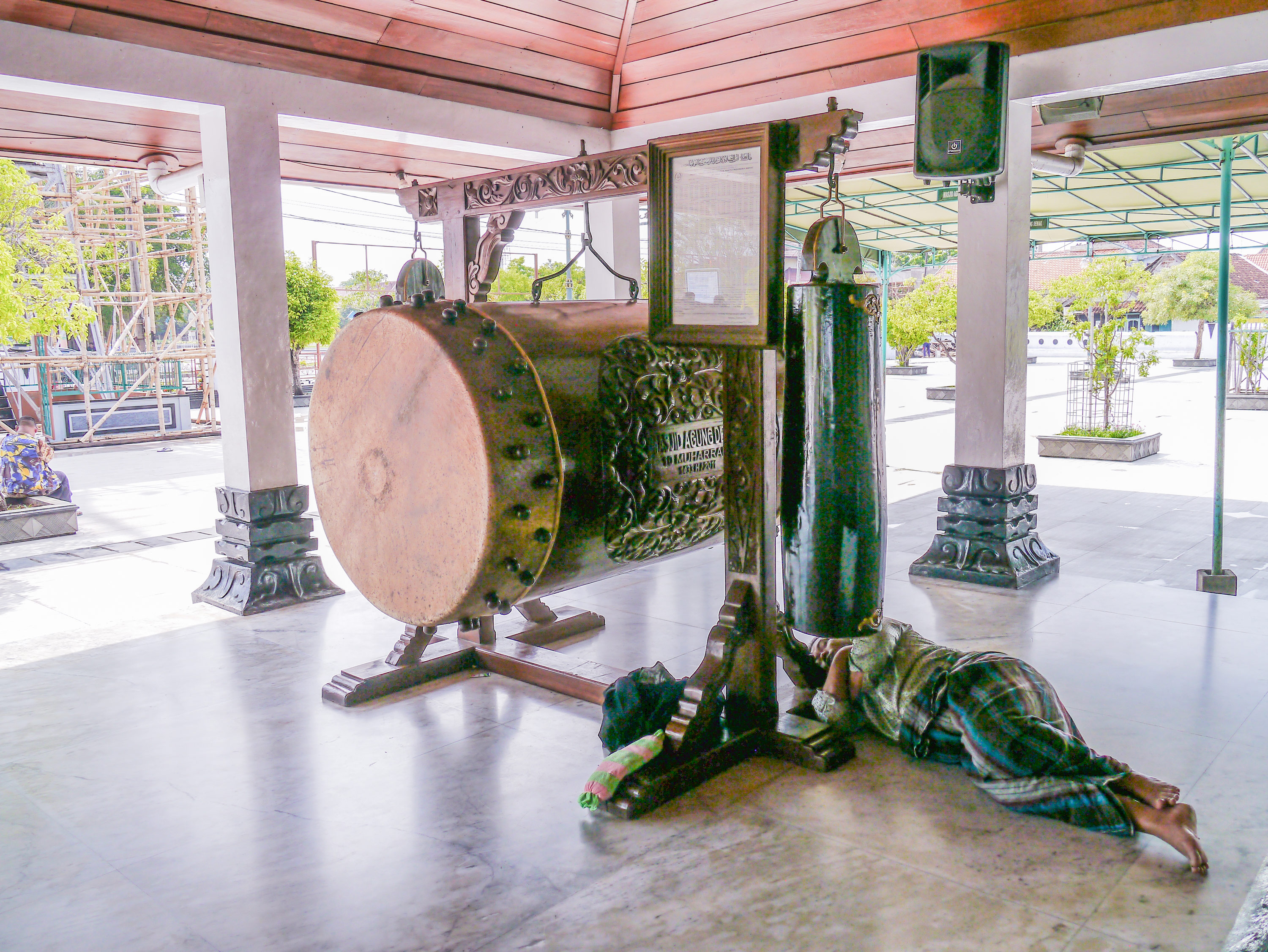 Large drum with two bells on either side held together by a wooden structure, seen in the same courtyard. A man is sleeping with his head on one of the wooden legs of the structure.