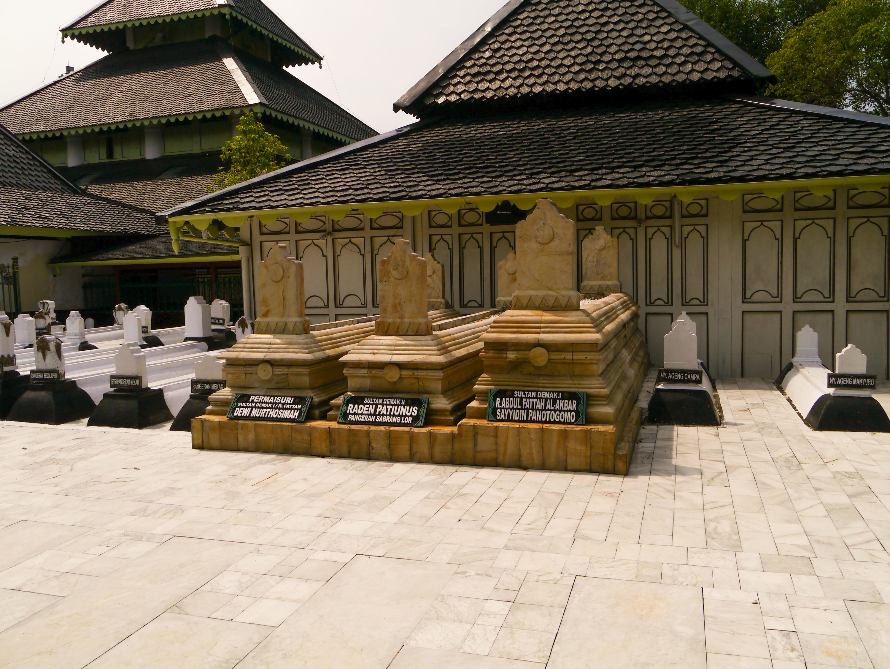 Three large sandstone/darker stone graves surrounded by smaller white graves in a courtyard with two and three-tiered buildings in the background.