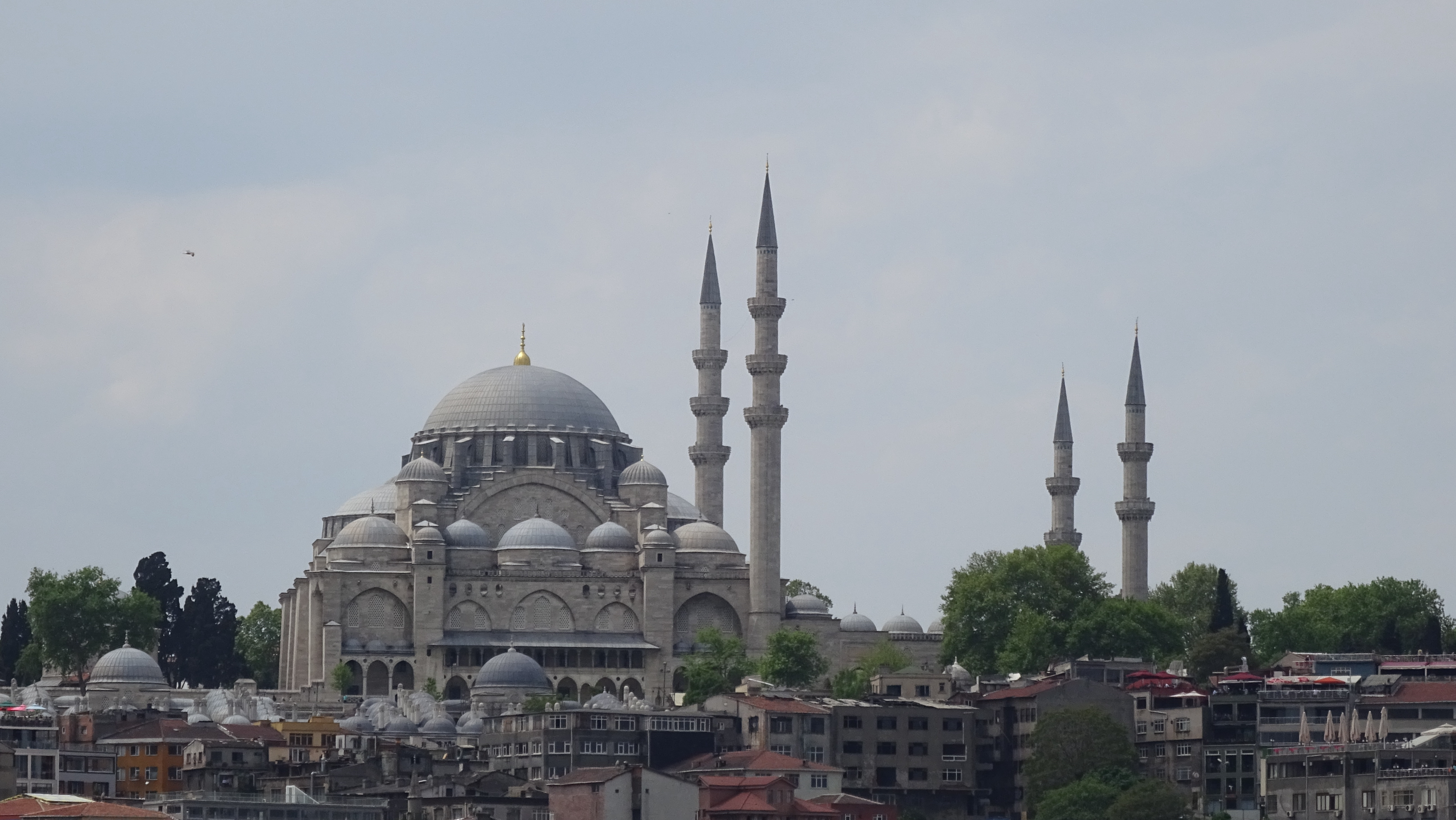 Grey stone mosque with darker grey domes and a golden finial along with four tall minarets to its right.