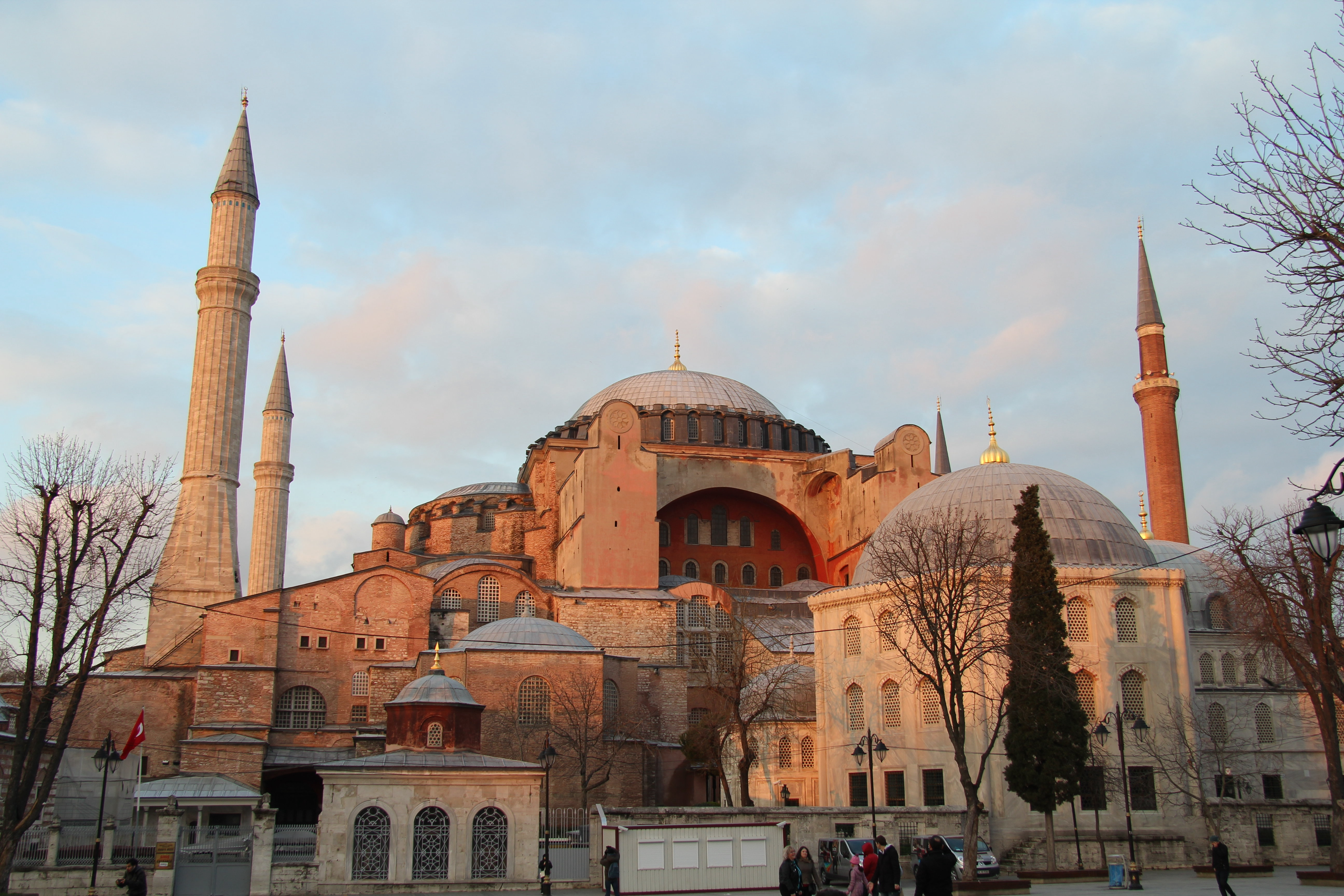 Sand-colored stone church/mosque complex with grey domes and four tall minarets around it. Golden finials are visible atop the domes and minarets.
