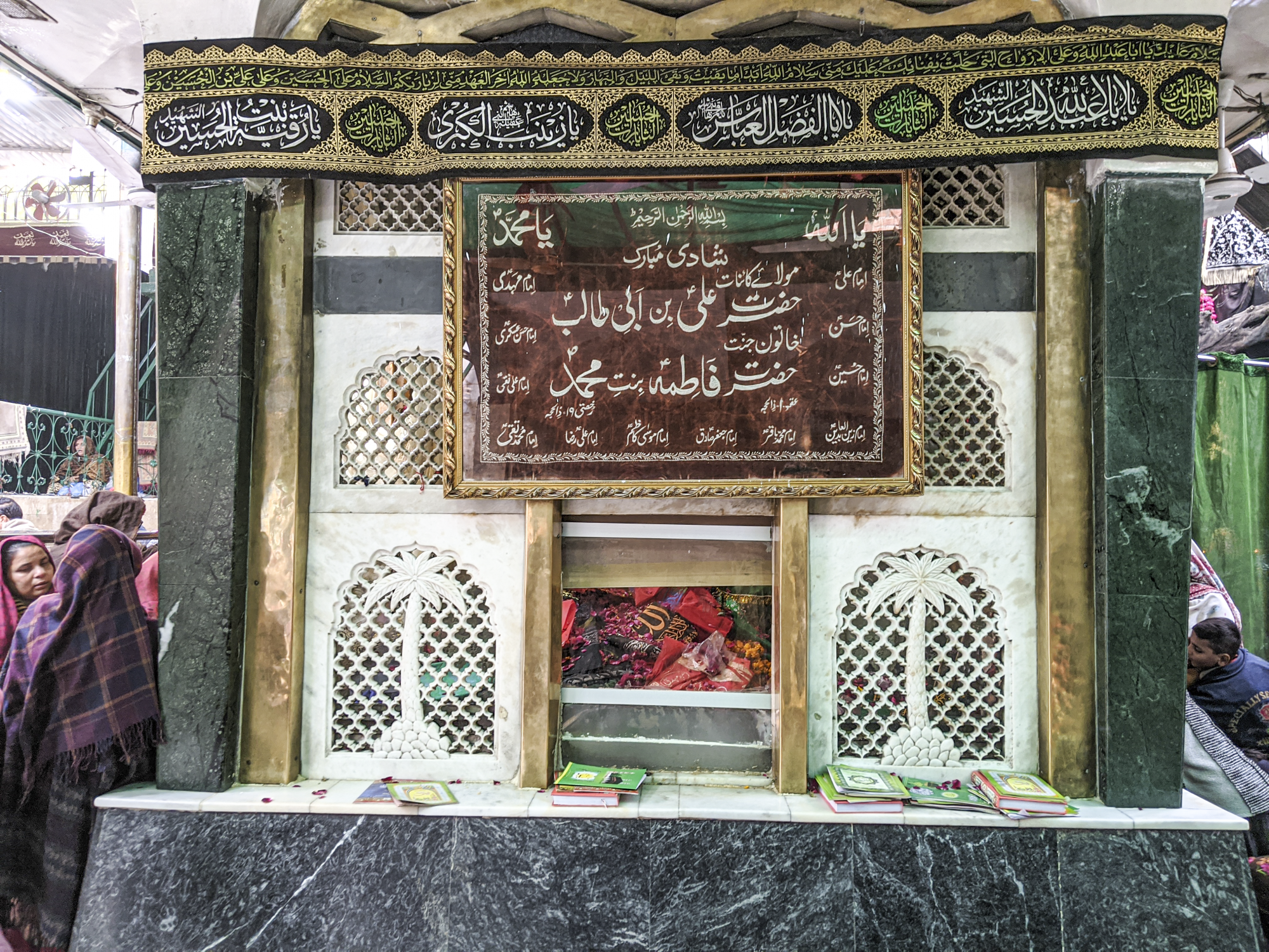 A shrine with Arabic calligraphy in black cartouches with white and green text lining the top. Another hanging brown banner has more Arabic text on it. A latticed white screen in six panels stands in the middle of the structure, with an engraved coconut tree in the center of each of the bottom panels.