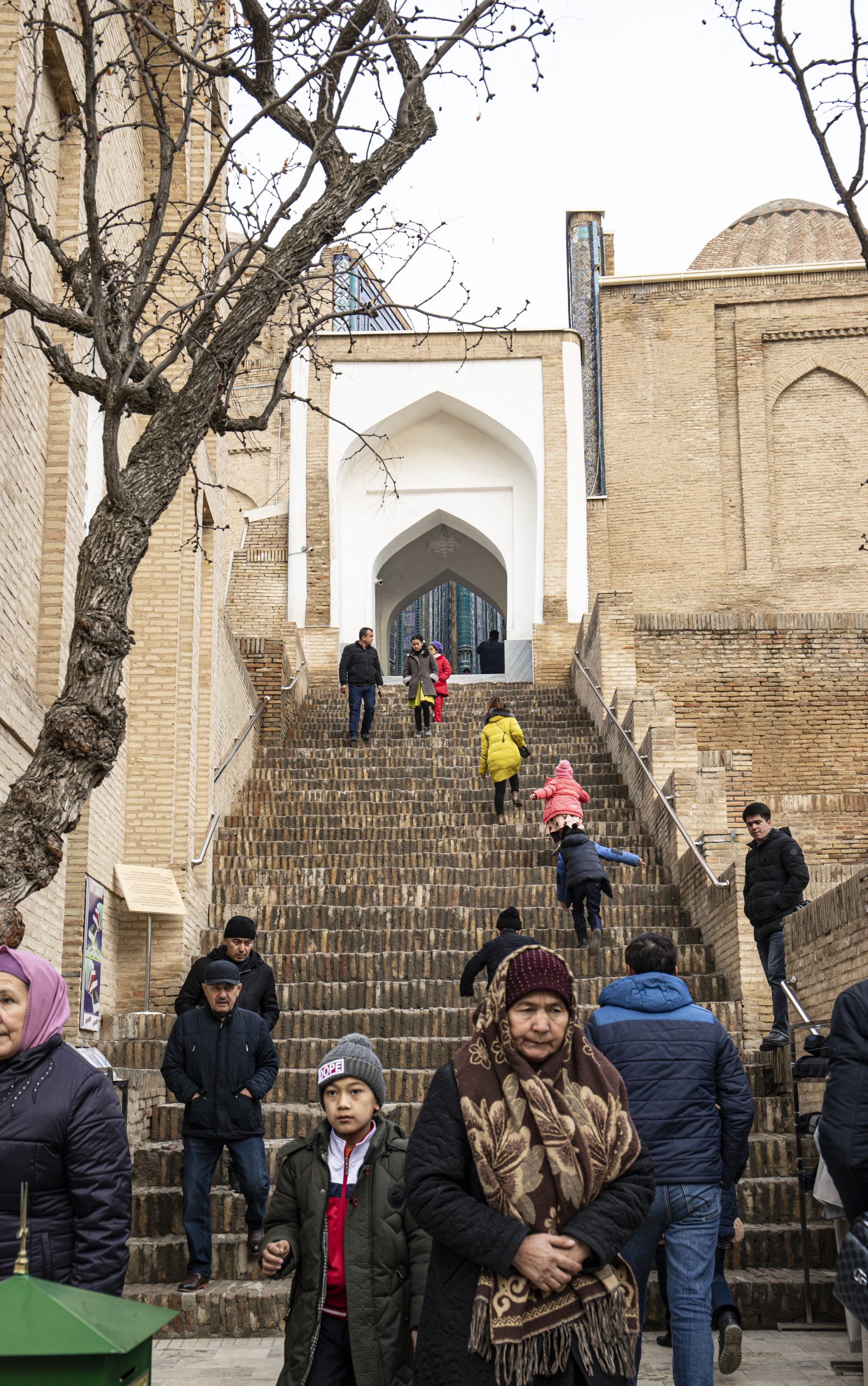 Stone staircase leading up to the entrance of a building. Various people can be seen going up and down the stairs, including brightly dressed children.