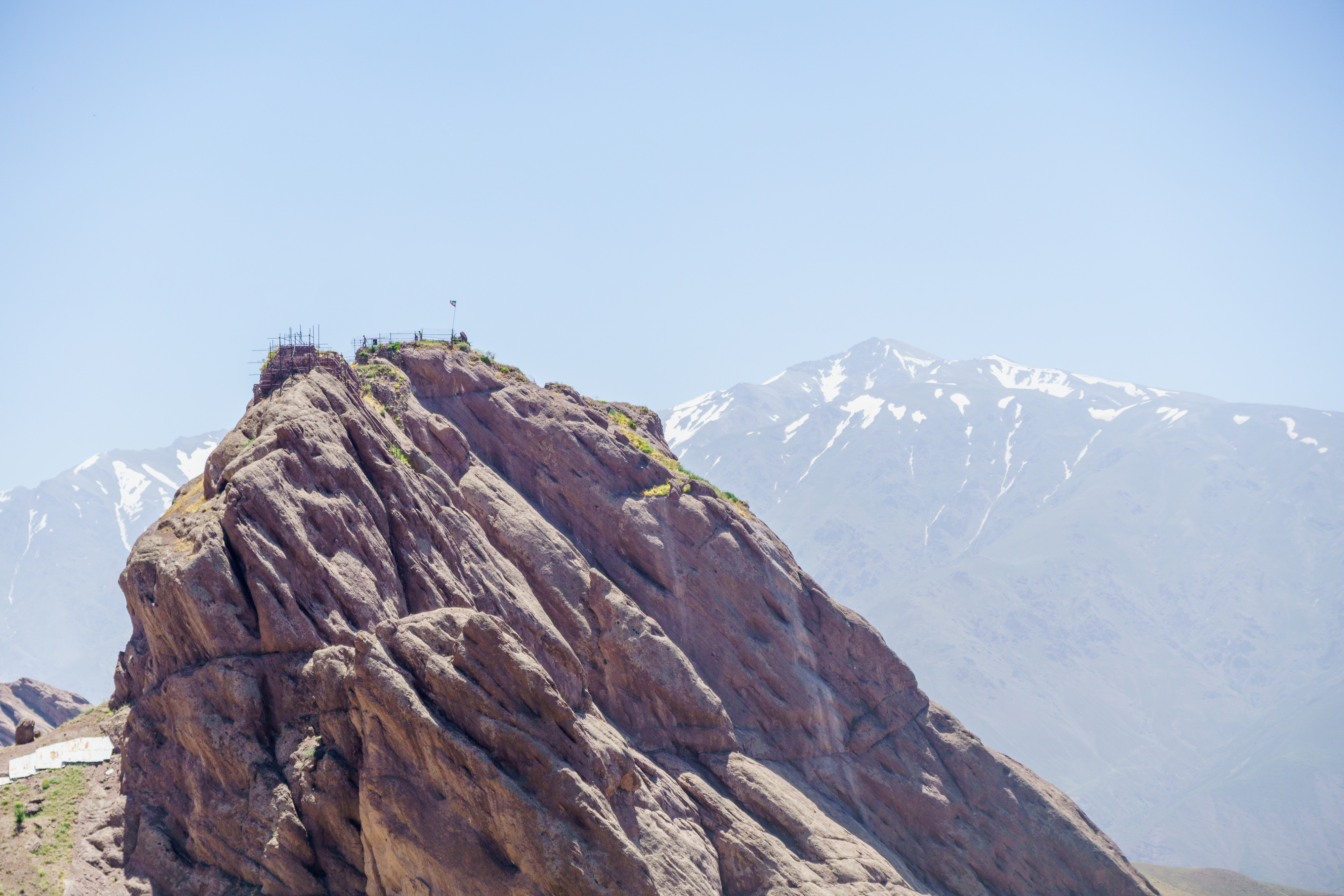 Photograph of a large rock with a small flag flying on its top. Snow-capped mountains are visible in the background.