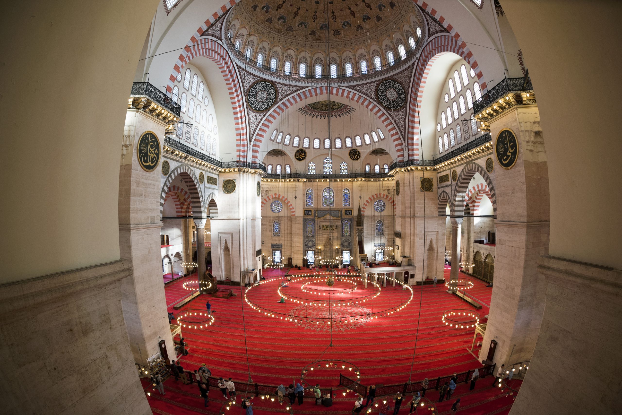 Fisheye view of the interior of the mosque which shows various chandeliers and red carpeted floors.
