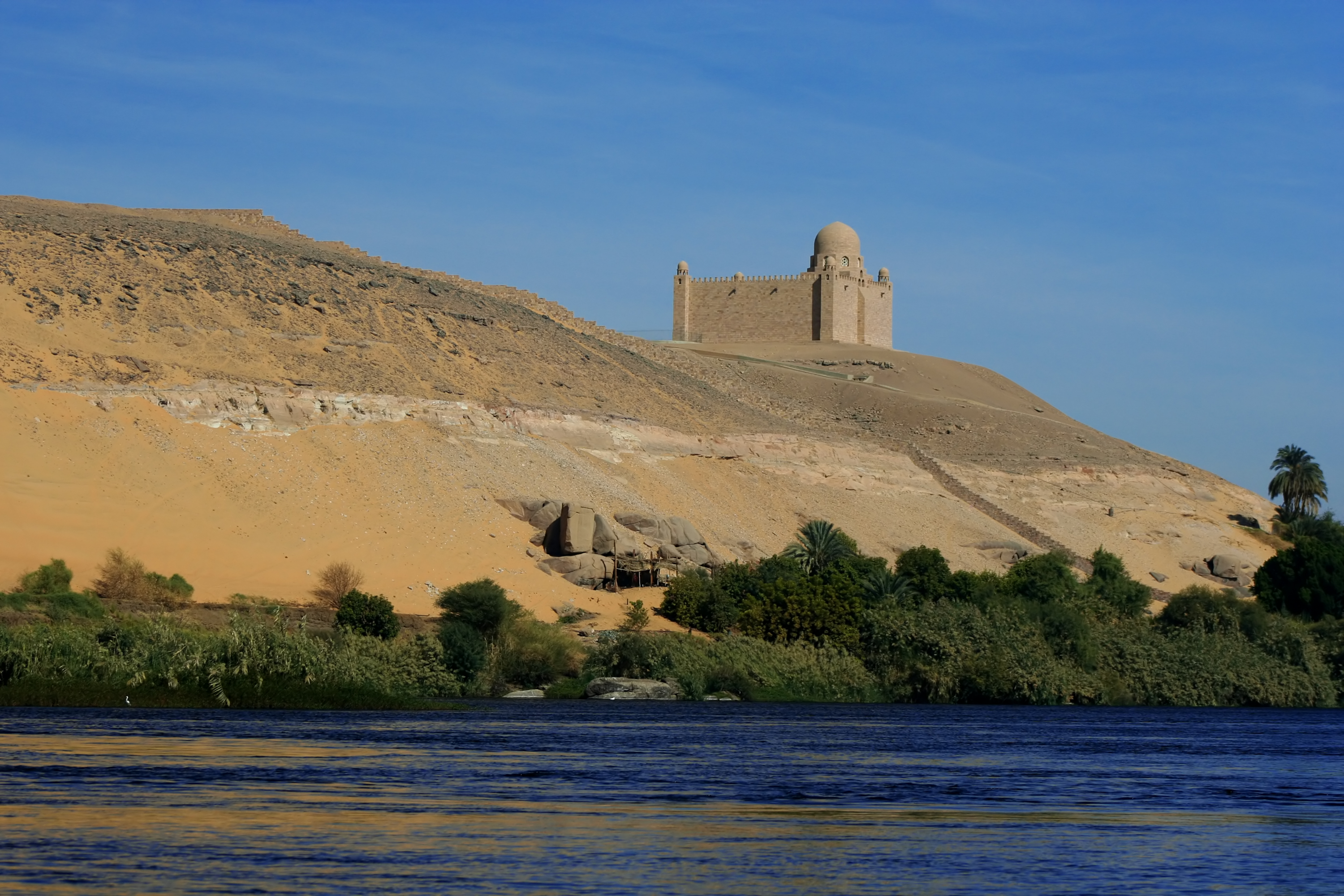 Domed mausoleum visible on a slight hill in the horizon. A river with greenery growing around it are seen in the foreground.